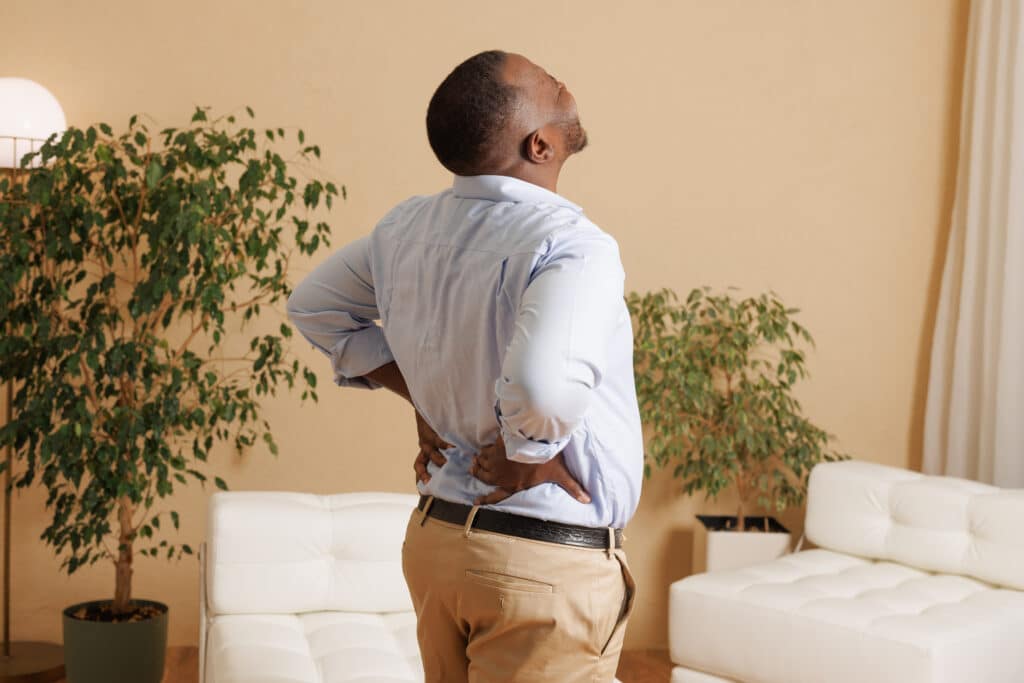 Man experiencing severe lower back pain from spondylolisthesis at home. he holds his back with both hands, grimacing in discomfort.